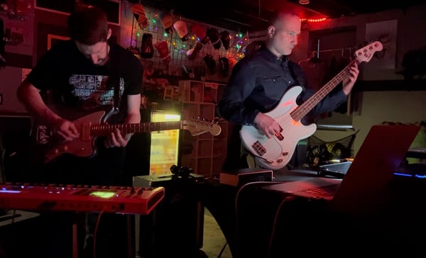Two men playing guitars in a dark room with ambient lighting standing in front of a table holding other music gear and a keyboard.