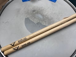 Two drum sticks resting on a snare drum. The brand of drum stick is Vater and the model is Super Jazz.