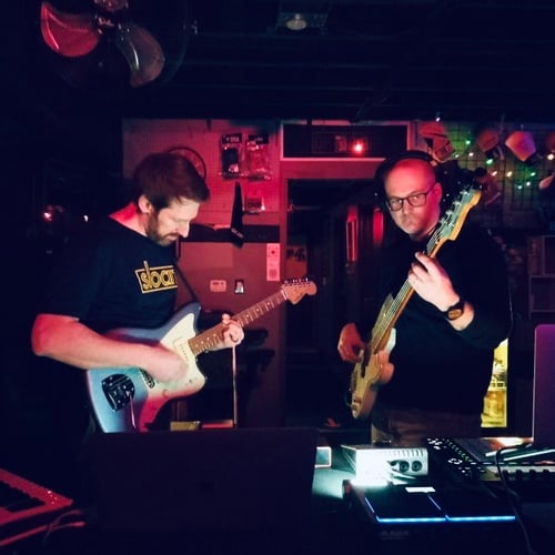 Two men playing guitars in a dark room with ambient lighting standing in front of a table holding other music gear.