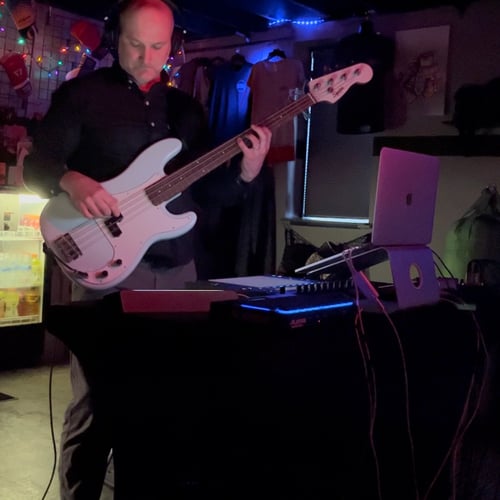 A man playing bass guitar in a dark room with ambient lighting standing in front of a table holding a computer and other music gear.