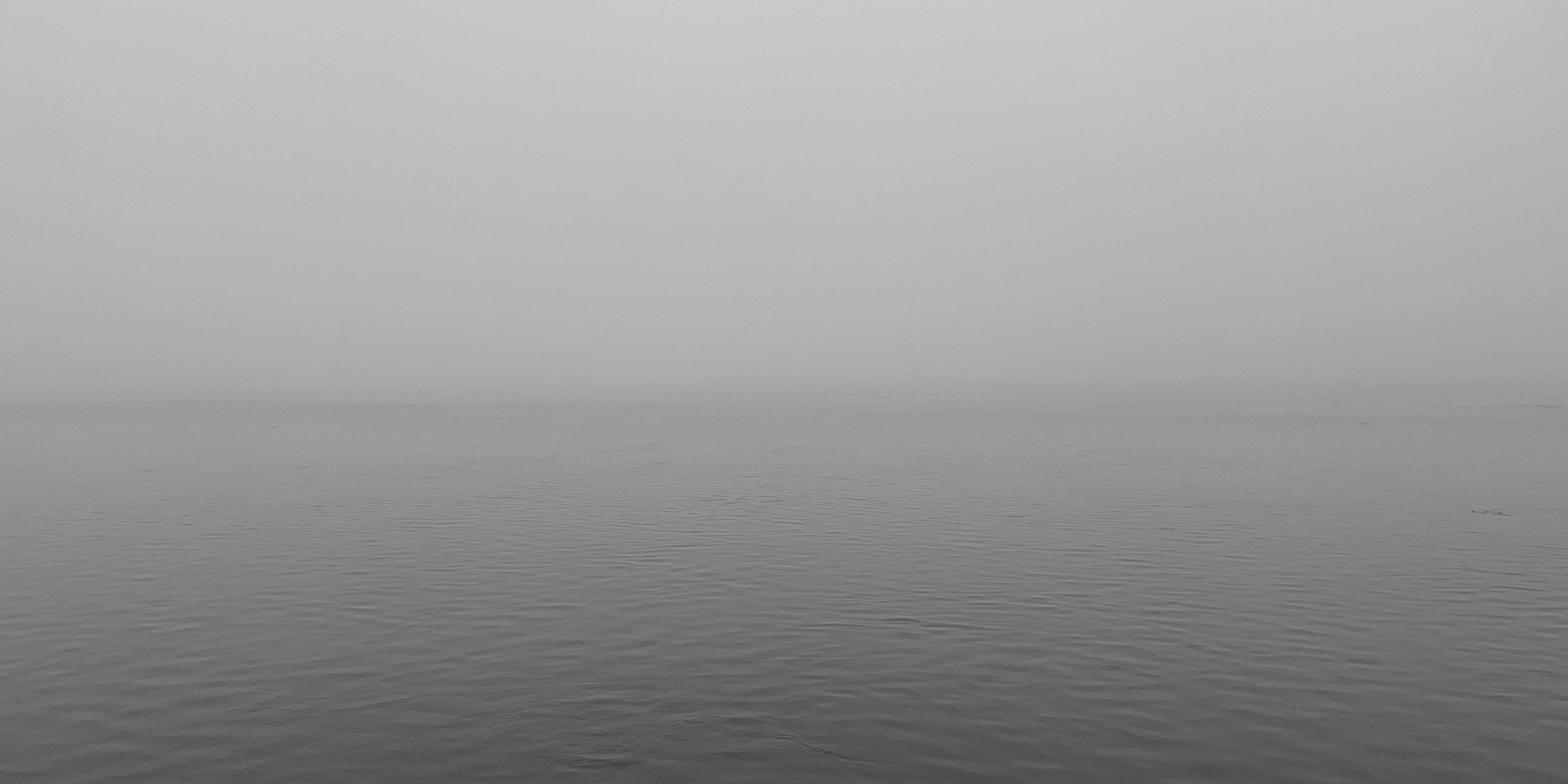 Black and white image of a calm lake with a blurry horizon behind fog under a gray sky