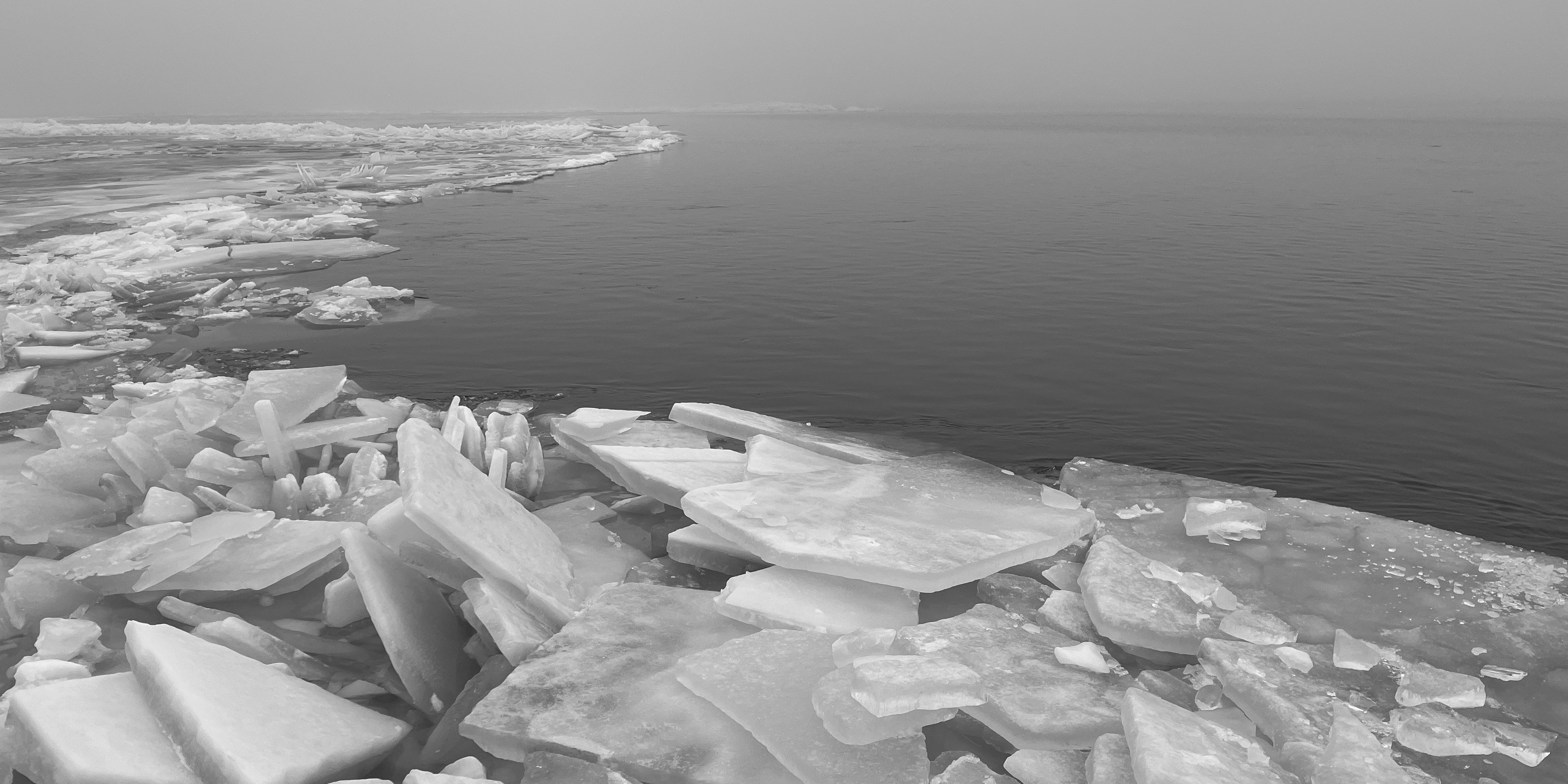 Black and white image of broken ice sheets building up at the edge of a calm lake under an overcast sky.