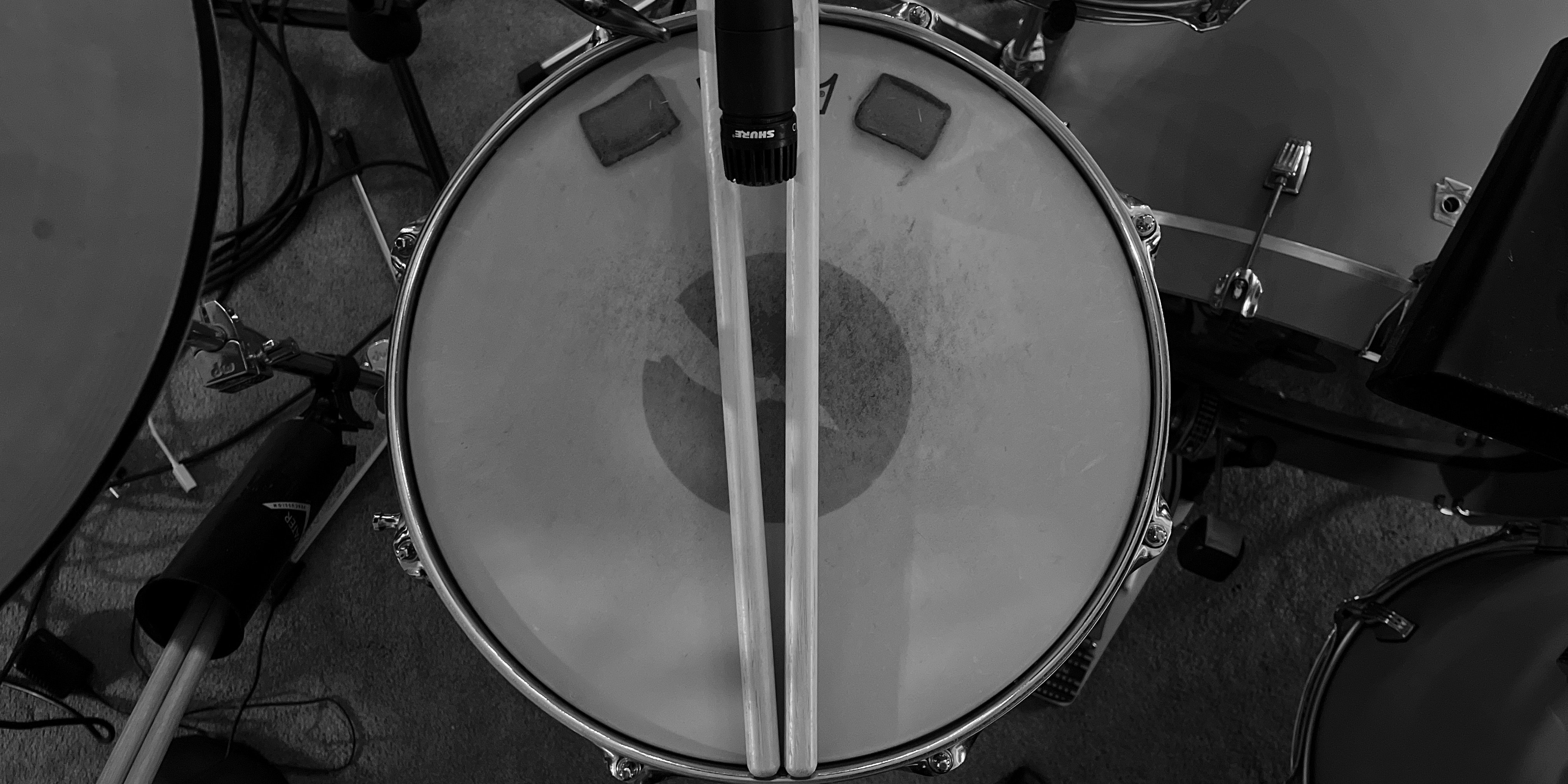 Aerial view of a drum set focused on the snare drum with two drumsticks resting on it and a microphone pointed at the center of the drumhead.