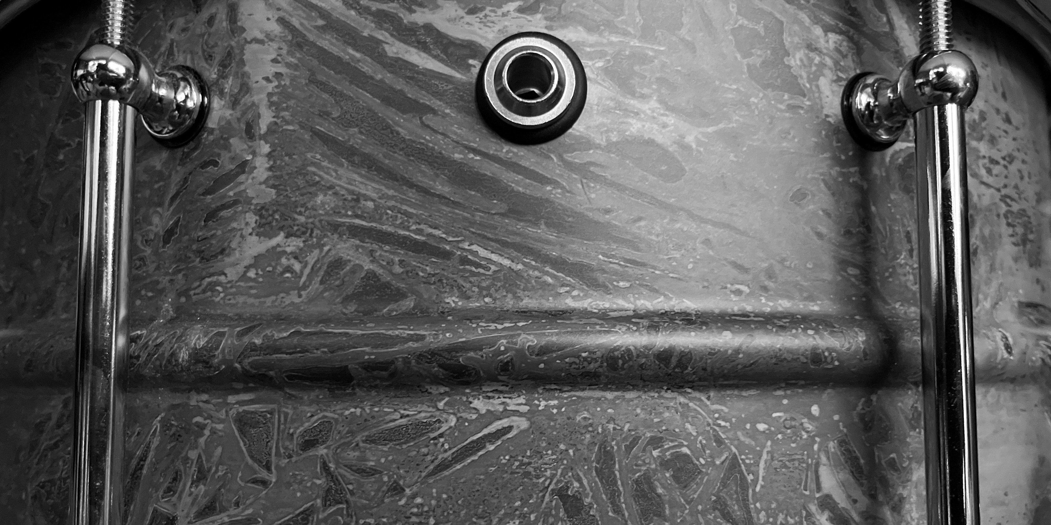 Closeup image of the side of a metal snare drum showing the pattern of the patina along with two lugs and the air vent centered in black and white