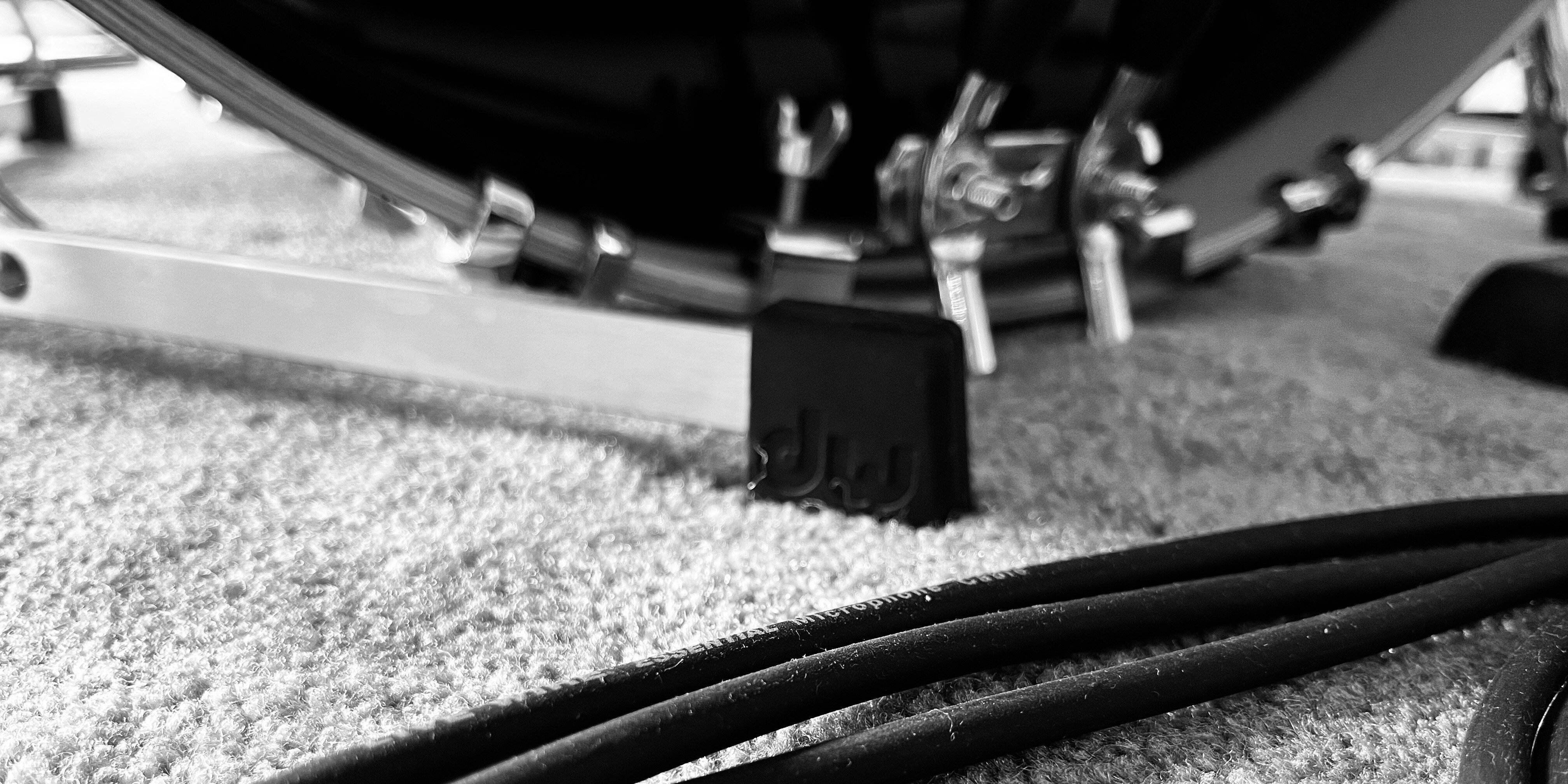 Black and white image taken at the base of a kick drum showing the leg of a cymbal stand and instrument cables in the foreground